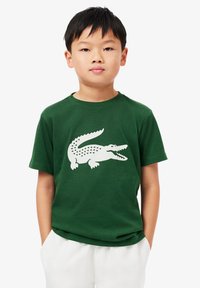 T-shirt en coton vert avec un grand graphique de crocodile blanc, manches courtes et col rond, porté avec un pantalon blanc.