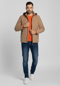 Chaqueta color marrón claro con cremallera al frente y capucha, con detalles en naranja, sobre un suéter naranja. Combinada con vaqueros oscuros y zapatillas blancas.