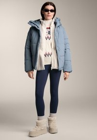 Világoskék steppelt dzseki, krémszínű pulóver piros és kék mintákkal, sötétkék léggings, bézs színű, szőrmével bélelt csizmák. A megjelenést napszemüveg teszi teljessé.