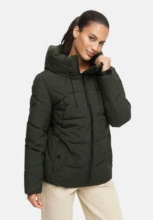 Vrouw met donker haar in een paardenstaart, gekleed in een donkergroene pufferjack met hoge kraag en beige broek, die met één hand de kraag vasthoudt.