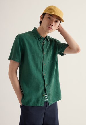 Junger Mann trägt grünes kurzärmeliges Hemd mit Knöpfen, gelbe Kappe, marineblaue Shorts und gestreiftes Unterhemd, posiert mit einer Hand hinter dem Kopf.