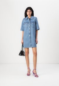 Τζιν φόρεμα με κοντά μανίκια, δύο τσέπες στο στήθος, κουμπιά μπροστά και ασύμμετρο στρίφωμα. Συνδυάζεται με μωβ ψηλοτάκουνα σανδάλια και μαύρη τσάντα.