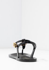 Svarta plastsandaler med platt sula, med en smal T-remdesign, guldbuckledetalj och en mjuk, glänsande yta.