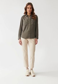 Camisa de color verde olivo con botones, dos bolsillos en el pecho, mangas largas y un discreto detalle en los hombros, combinada con pantalones beige y zapatillas blancas.
