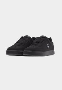 Paire de baskets basses noires avec des lacets noirs, des semelles plates, et un logo blanc "CK" sur le côté et "Calvin Klein" sur la languette.