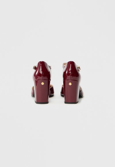 Chaussures à talons en cuir verni bordeaux avec un talon carré, une bride de cheville et des détails en métal doré à l'arrière. Texture lisse et brillante.