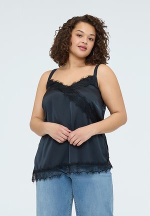 Femme aux cheveux bouclés portant une camisole noire à bordure en dentelle et un jean bleu clair, debout devant un fond gris clair uni.