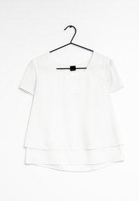 Vit kortärmad blus med rundad halsringning, lagerdesign och en slät textur. Hängande på en svart plastgalge mot en vit bakgrund.