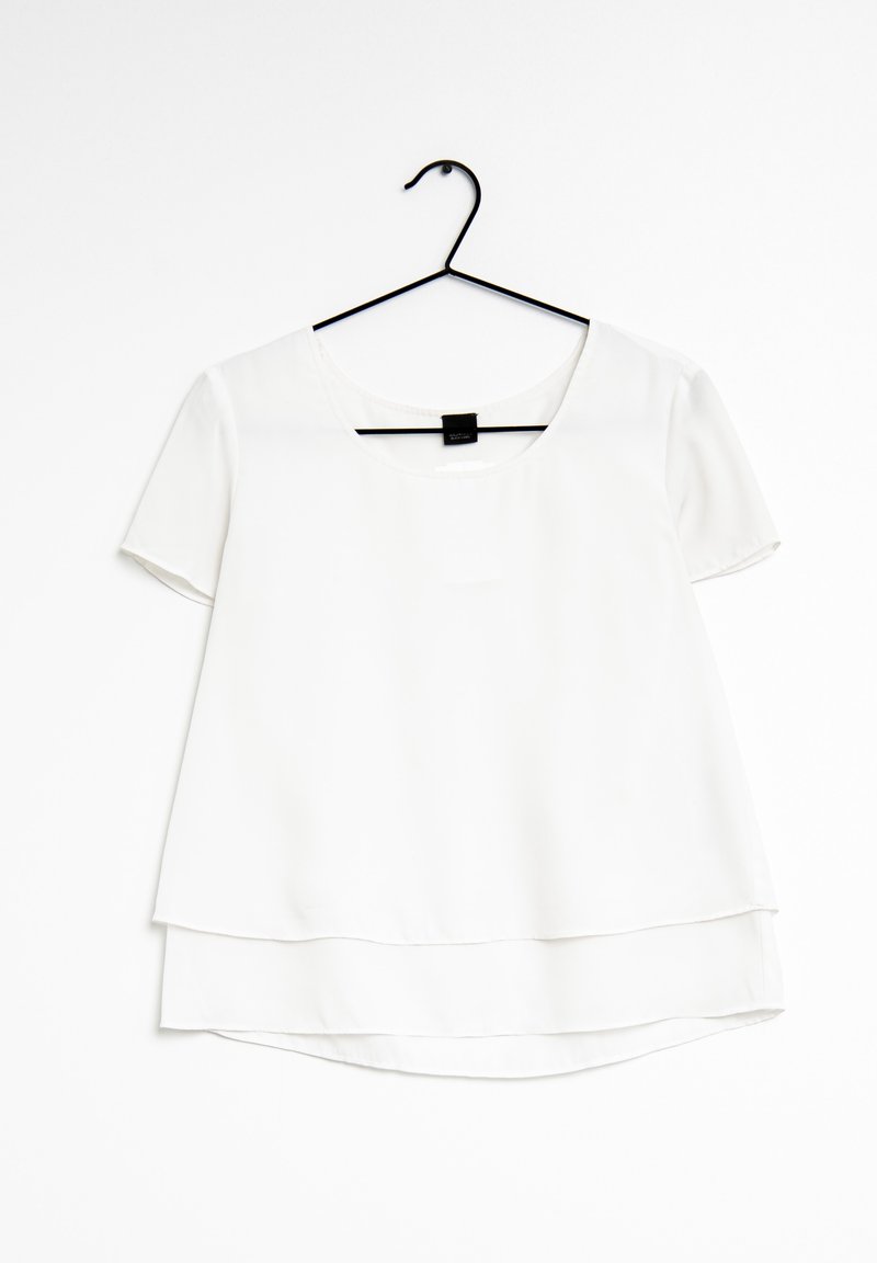 Vit kortärmad blus med rundad halsringning, lagerdesign och en slät textur. Hängande på en svart plastgalge mot en vit bakgrund.
