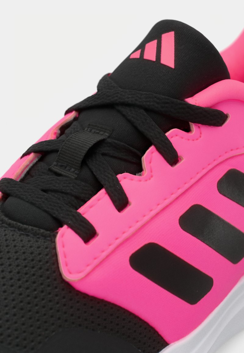 Zapato deportivo negro con superposiciones sintéticas rosas, tejido negro texturizado, cordones negros y tres rayas negras en el lateral.