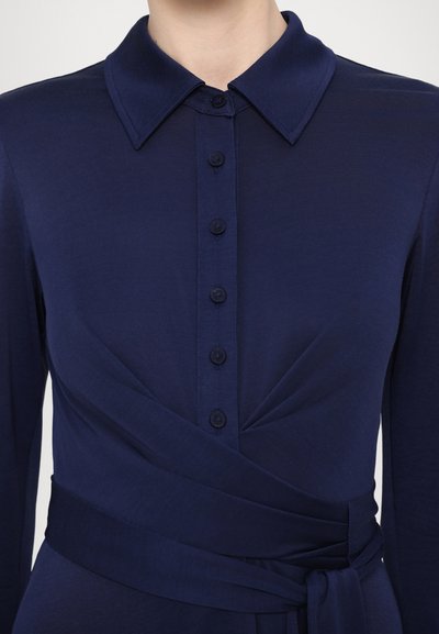 Tamnoplava haljina s okruglim ovratnikom na kopčanje, s dizajnom umotavanja i pojasom koji se veže u struku. Glatka tekstura s blagim sjajem i šest dugmadi u istom tonu.