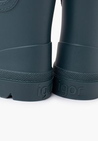 Bottes de pluie en caoutchouc vert foncé avec des semelles à grip texturé, détail de logo circulaire sur la tige et nom de marque embossé sur le côté.