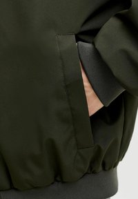 Grøn bomberjakke med glat, vandafvisende stof, ribbet manchet og en sidelomme integreret i designet.