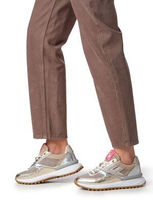 Chaussures de sport argentées et beiges avec des accents en mesh et métalliques, dotées d'une semelle texturée et d'un logo rose sur la languette. Portées avec un pantalon marron.