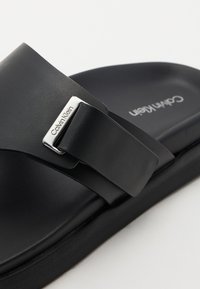Svart slide-sandal med en bred stropp, med en sølvlogo-spenne. Sålen er flat og teksturert, laget av syntetisk materiale.