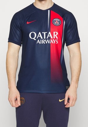 Une personne porte une tenue de football bleu marine du Paris Saint-Germain, avec une bande rouge sur le maillot, l’écusson du club, le swoosh Nike et le sponsor Qatar Airways.