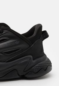Black athletic shoe featuring a textured sole, padded collar, and fabric upper with mesh cutouts and branding detail on the side.