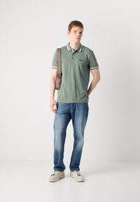 Žalias polo marškinėliai su baltais dryželiais ant apykaklės, logotipas ant krūtinės, derinami su mėlynomis džinsais ir smėlio spalvos sportbačiais. Modelis laiko rudą petinę krepšį.