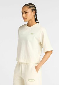 Vrouw met gevlochten haar in een crèmekleurig New Balance Athletics T-shirt met korte mouwen en bijpassende joggingbroek, met handen in de zakken, kijkend naar de zijkant.