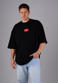 Mann trägt ein übergroßes schwarzes T-Shirt mit rotem Logo und helle blaue Jeans, posiert mit einer Hand in der Tasche vor einem grauen Hintergrund.