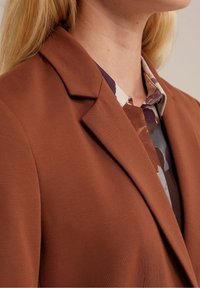 Brun kavaj med notch-krage och slät textur, i kombination med en mönstrad skjorta med dämpade färger och abstrakta mönster.