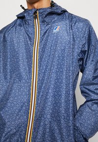 Blå mønstret vindjakke med lynlås foran, gule, orange og sorte striber, justerbar hætte og letvægtigt materiale.