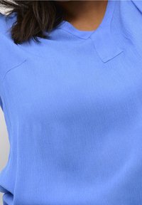 Top azul claro de manga corta con una tela texturizada, que presenta un sutil escote en V y un mínimo detalle de costura. Diseño simple y limpio.
