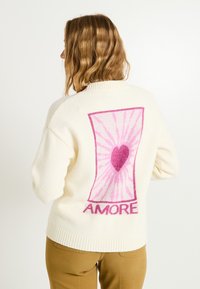 Pull crème avec une ourlet côtelé, mettant en avant un grand graphique rose représentant un cœur et des rayons, sur lequel est écrit "AMORE" au dos.