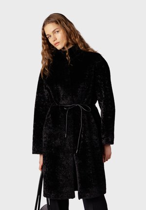 Femme aux cheveux ondulés portant un manteau noir texturé ceinturé tenant un sac à main noir sur un fond clair uni.
