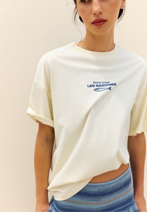 Femme portant un t-shirt blanc avec "Saveur unique LES SARDINES" et un graphisme de poisson, associé à une jupe rayée bleue.