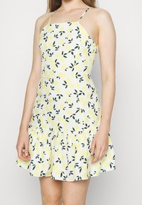 Robe à motifs de citrons avec des bretelles spaghetti, présentant un corsage ajusté et un ourlet à volants. Principalement blanche avec des accents jaunes et verts.