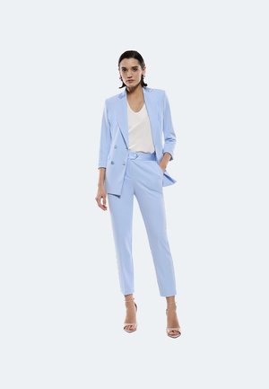 Suit su misura azzurro chiaro con giacca doppiopetto e pantaloni slim-fit. Texture liscia, abbinato a un top bianco e tacchi nudi con cinturini.