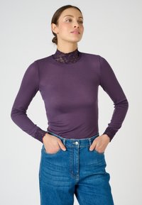 Top viola a maniche lunghe con colletto alto in pizzo e dettagli in pizzo sui polsini, abbinato a jeans in denim blu a vita alta.