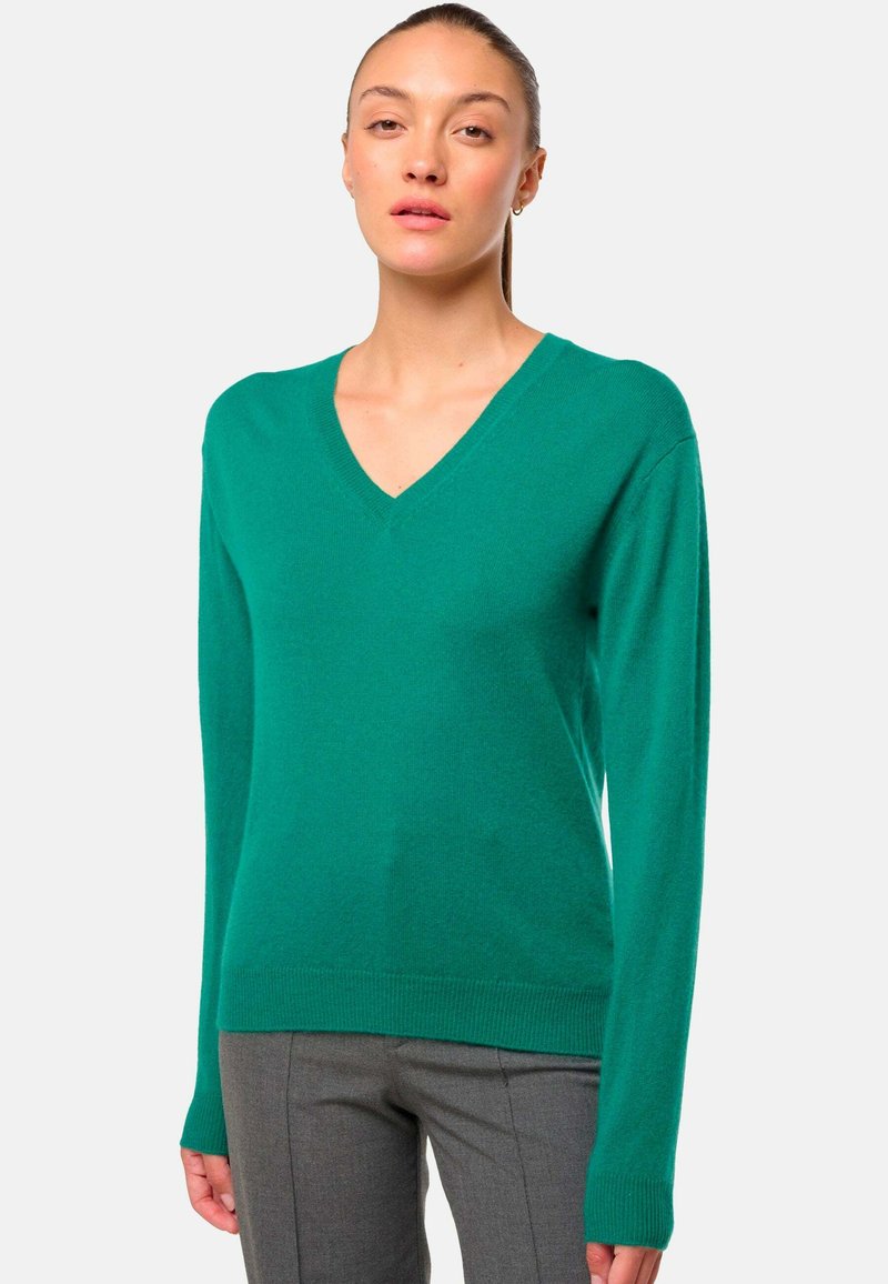 Pull vert en V en tissu tricoté doux, avec des manches longues et un ourlet côtelé. Porté avec un pantalon gris sur mesure, mettant en valeur la texture du pull.