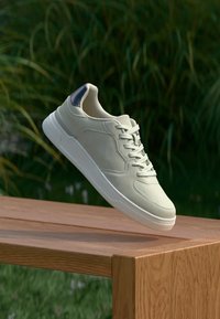 Helle beige Sneaker mit glatter Textur, Schnürdesign und einem kontrastierenden dunkelblauen Fersenakzent. Auf einer Holzoberfläche positioniert.