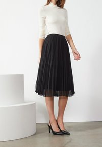 Gonna nera plissettata con sovrapposizione trasparente e vita elastica, abbinata a décolleté nere a punta. Top bianco con maniche lunghe. Sfondo minimalista.