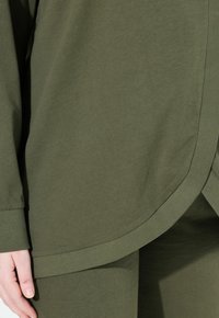 Primer plano de una persona que lleva una camiseta de manga larga y pantalones de color verde oliva, con enfoque en los detalles de los dobladillos curvados y los puños cerca de la mano izquierda.