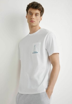 Młody mężczyzna z krótkimi brązowymi włosami, ubrany w białą koszulkę z małą grafiką żaglówki na kieszonce na piersi.