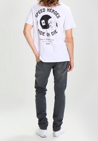 Camiseta blanca con gráfico negro de un cráneo y un casco, con el texto "Speed Heroes Ride or Die." Un par de jeans oscuros y zapatillas blancas.