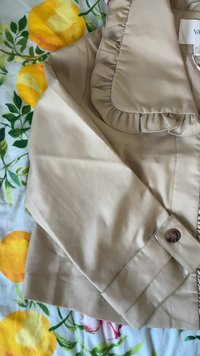 Jachetă bej cu mâneci lungi, detaliu cu guler fronsat și manșete cu nasturi, așezată pe o pătură florală cu fructe galbene și frunze verzi.