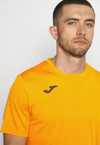 Svetlo oranžové športové tričko s krátkymi rukávmi, vyrobené z hladkej látky. Na ľavej hrudi má čierne logo.