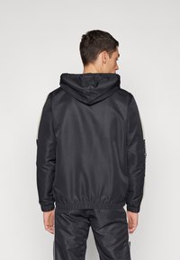 Chaqueta con capucha en negro, que presenta una cintura ajustada, textura suave y acentos en contraste. La vista trasera destaca el diseño minimalista.