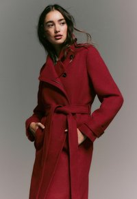 Red wool coat with a belted waist, wide lapels, and button accents. Features pockets and a relaxed fit with long sleeves.