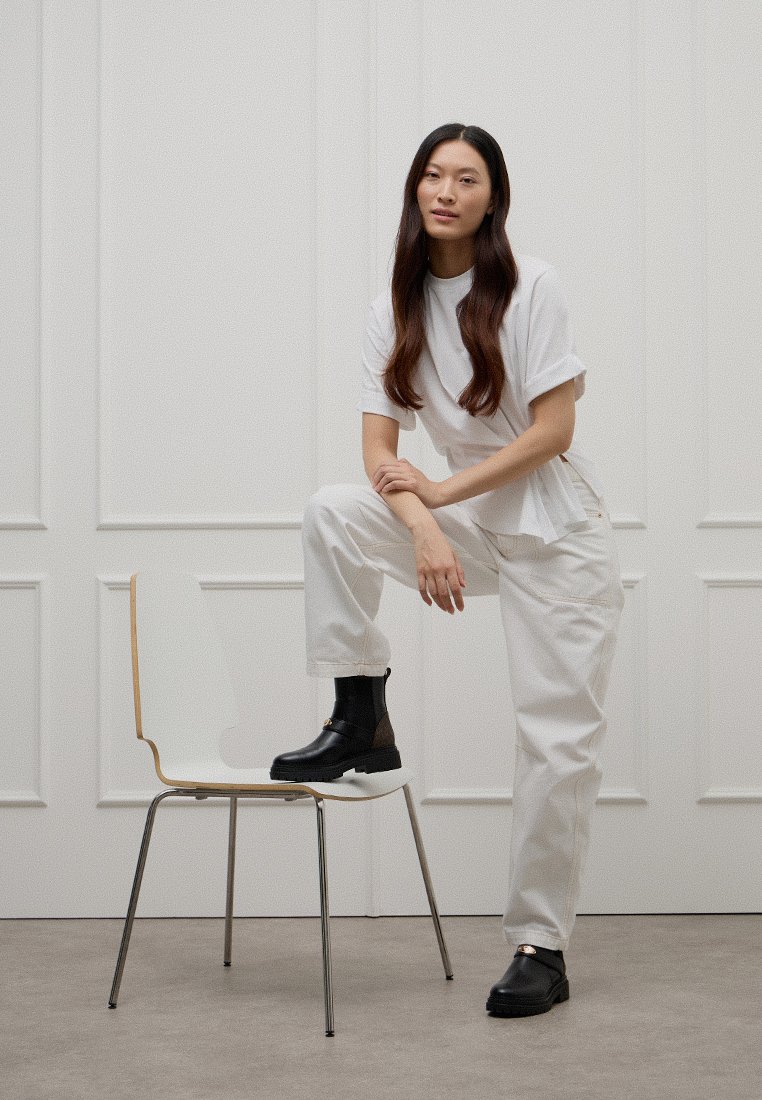 Młoda osoba z długimi włosami, ubrana w białe ubrania i czarne botki, pozująca z jedną nogą na białym krześle przy białej panelowej ścianie.