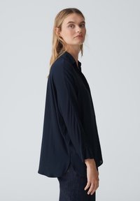 Blusa a maniche lunghe blu navy con colletto, fessure laterali e una texture liscia, caratterizzata da una vestibilità rilassata e un orlo leggermente arrotondato.