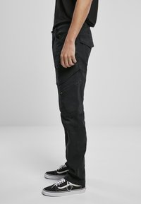 Schwarze Cargo-Hosen mit slim fit, ausgestattet mit mehreren Seitentaschen und einem strukturierten Stoff. Getragen mit schwarzen Sneakers und einem schwarzen T-Shirt.