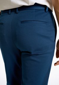 Marinefarbene Hose mit glatter Textur, maßgeschneidertem Design, zwei Gesäßtaschen und schlanker Passform. sichtbarer Bund mit Gürtelschlaufen.