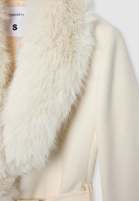 Cappotto color crema con un morbido colletto in pelliccia sintetica, realizzato in tessuto testurizzato. Presenta una cintura e un'etichetta con la scritta 'S' per la taglia.