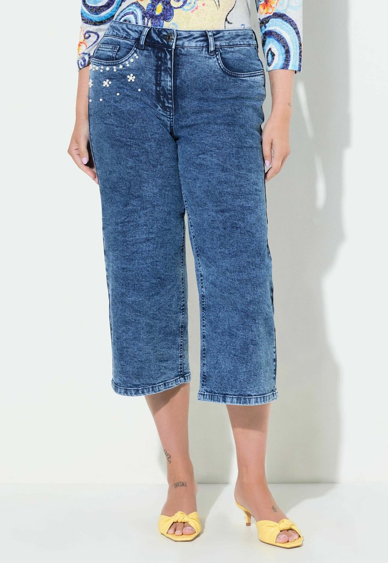 Kovové kalhoty v středně modré barvě s vybledlým ombre efektem, zdobené květinovými aplikacemi na levé kapse a rovnými nohavicemi.