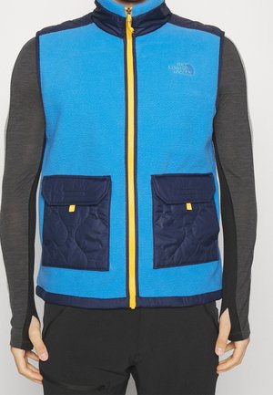 Blauwe fleecevest met contrasterende marineblauwe zakken en gele rits, gedragen over een donkergrijze top met lange mouwen, middelstuk zichtbaar.